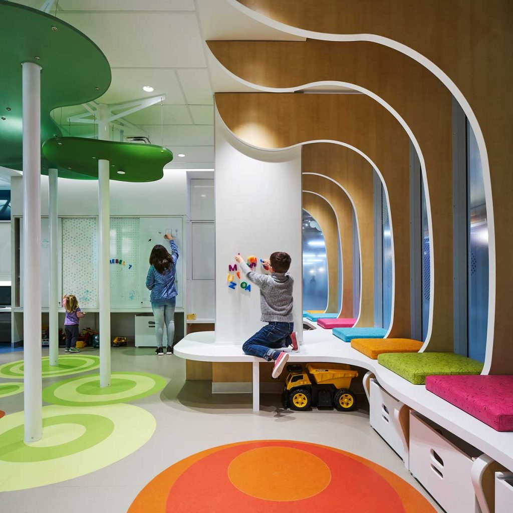 A boy plays with letters on a magnetic wall at the SickKids PlayPark with curved wall columns that resemble trees. Support beams are surounded by green organic forms at their tops to resemble trees with leaves.