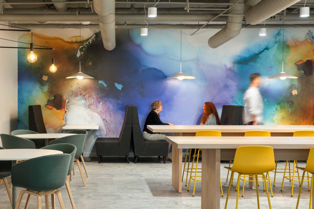 The employee cafe area at the Cooperators Regina has a watercolour inspired accent wall, booths, four top tables as well as long harvest tables.
