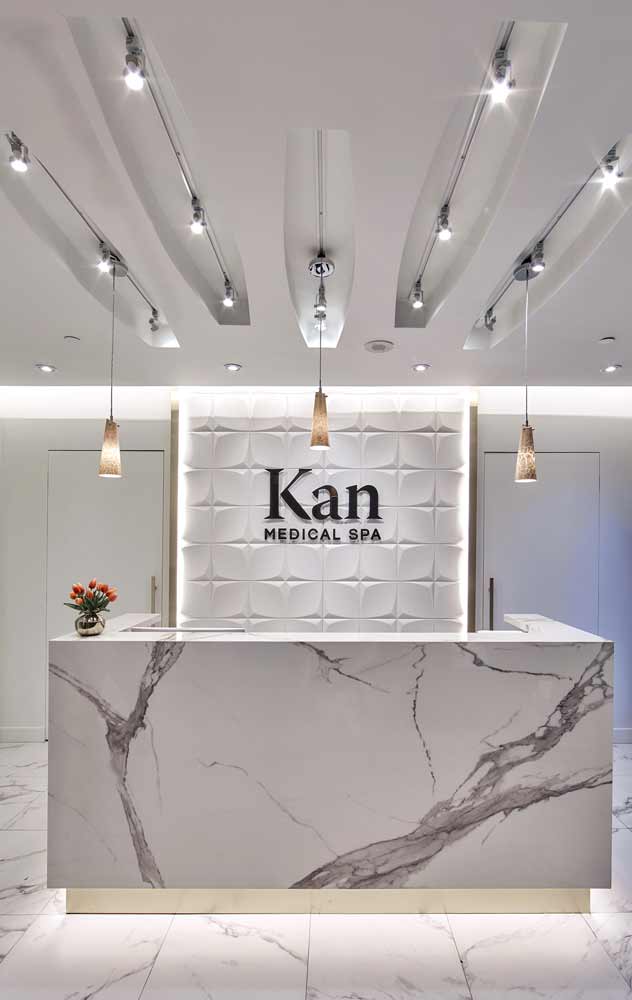 Reception desk in white marble with gray veining, the geometric textured feature wall behind in, and golden accents in light pendants above the desk