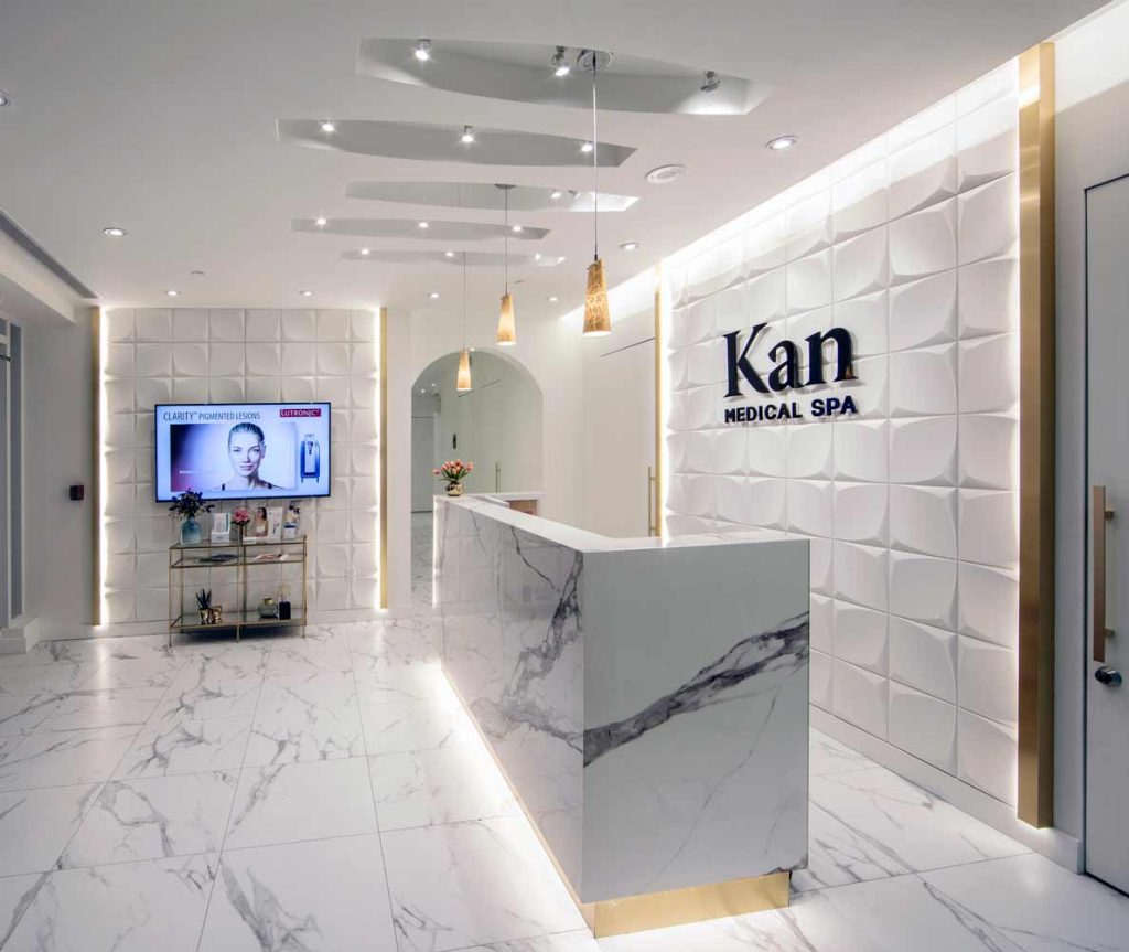 Entire reception area in the contemporary white scheme with golden accents aroun the geometric feature walls and below the reception desk, as well as lighting. A white wall hallway leading to exam rooms is visible in the background.