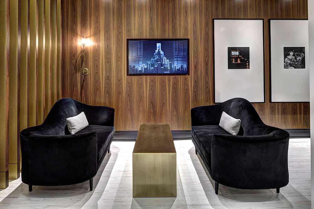 Lounge area with two black velvet sofas facing each other against a wood finish wall with oversized lifestyle photographs and a built-in digital screen