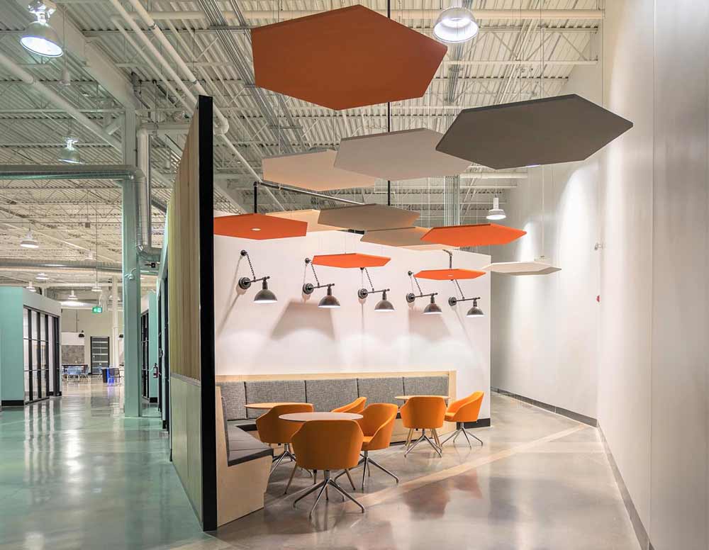 A more secluded collaboration area with bench seating and chairs and tables with orange accents in furniture and hanging hexagon details above