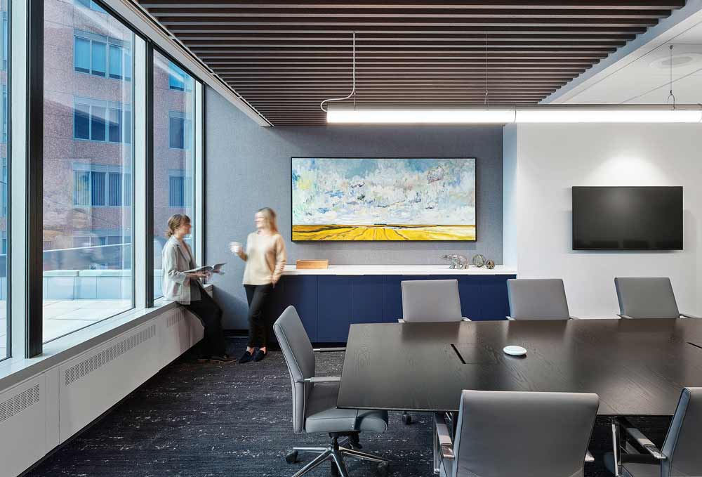 Light filled corner of large meeting room featuring a large landscape painting in yellows and blues above a dark blue built in cabinet.