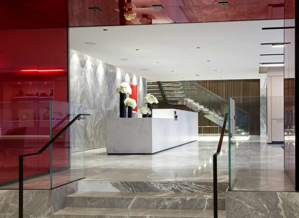 A view from the building entrance through a red accent wall feature toward the White marble reception desk on the ground floor with an impressive stair case right beside it leading upstairs