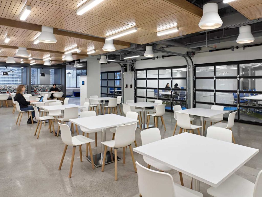 A view of the bright and open cafeteria space with lots of tables and seating, with adjacent meeting rooms behind glass 'garage doors'.
