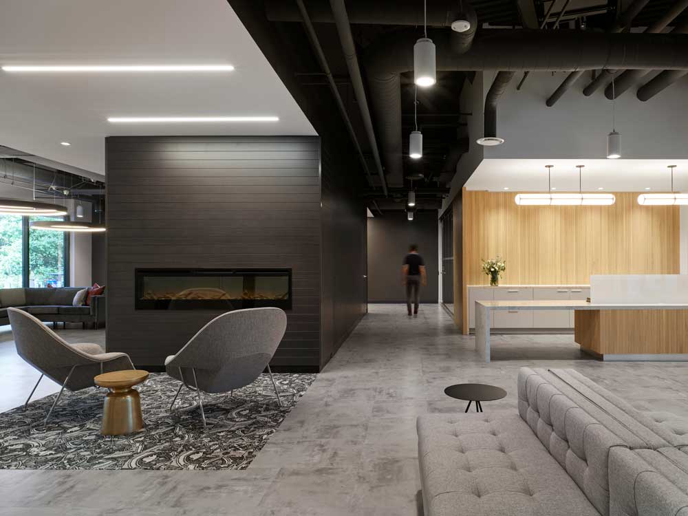 Central lounge area by a feature grey brick fire place next to a reception area. Mix of wood, marble, black paint and accents, exposed piping in the ceiling gives this interior a contemporary industrial yet inviting look