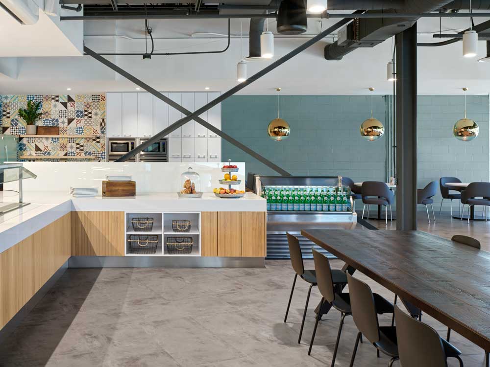 A broad view of the cafeteria. Wooden cabinetry and crisp white counters, colourful tile wall and plentry of tables and seating below golden globe lighting pendants