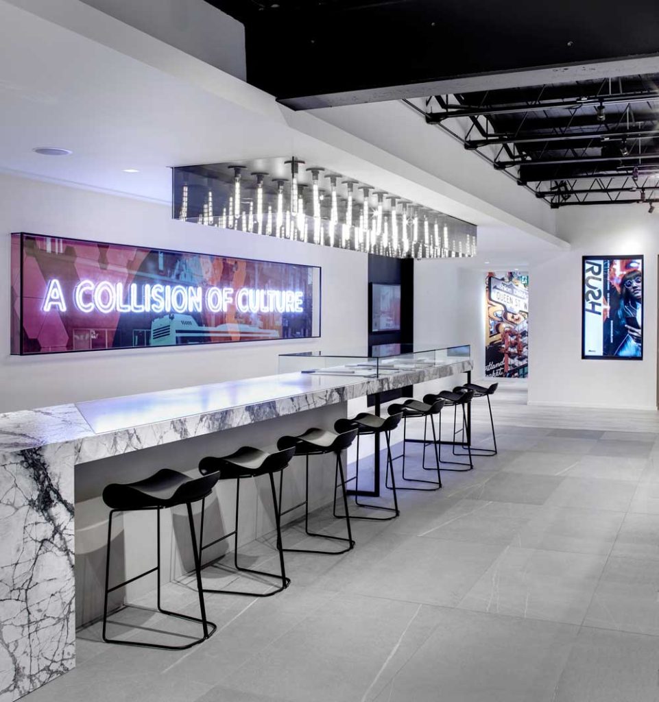 Pops of neon, with a punchy ‘A Collision of Culture’ sign slogan and in front of it counter with marketing materials and statement lighting above, with pops of colorful art in the background