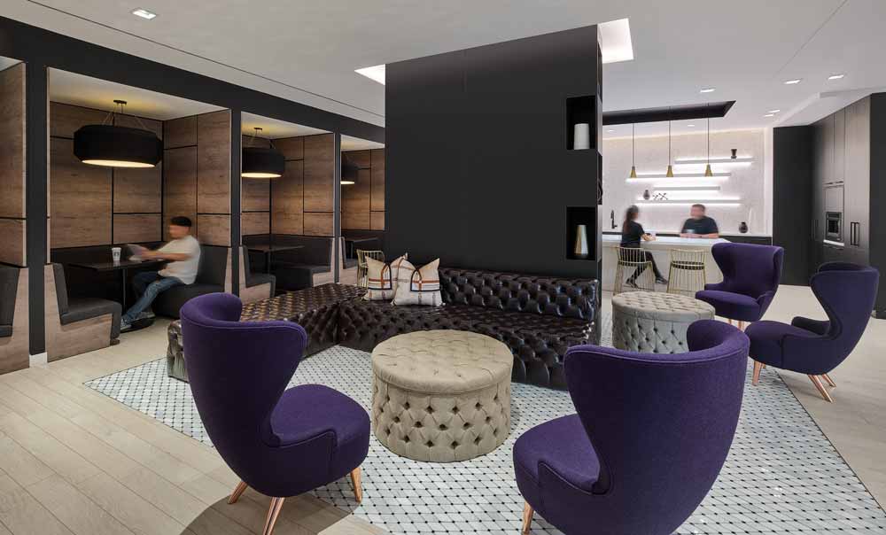 An overall view of the party room lounge area with purple accent chairs and booths in the foreground and the kitchen area peeking through in the background