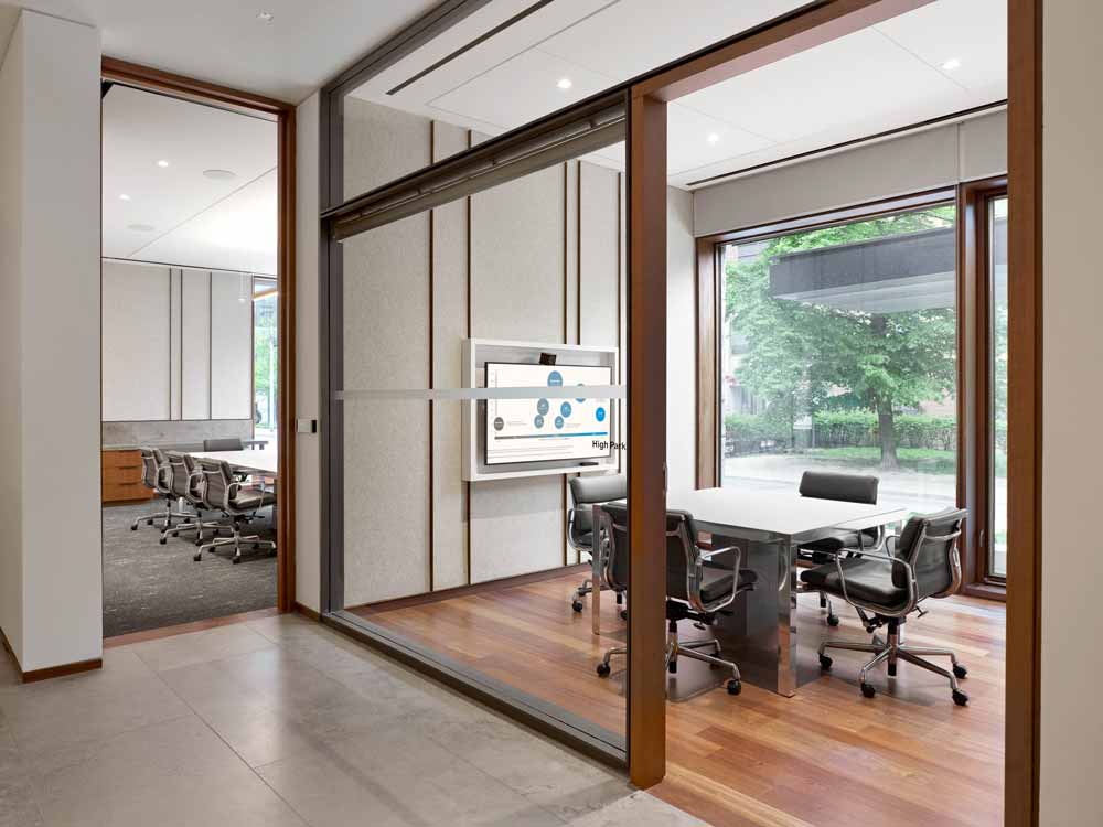 A view into two adjacent meeting rooms through a glass wall, where modern decor and interiors are warmed up by the use of wooden finishes on floors, door frames and wood slat details on walls