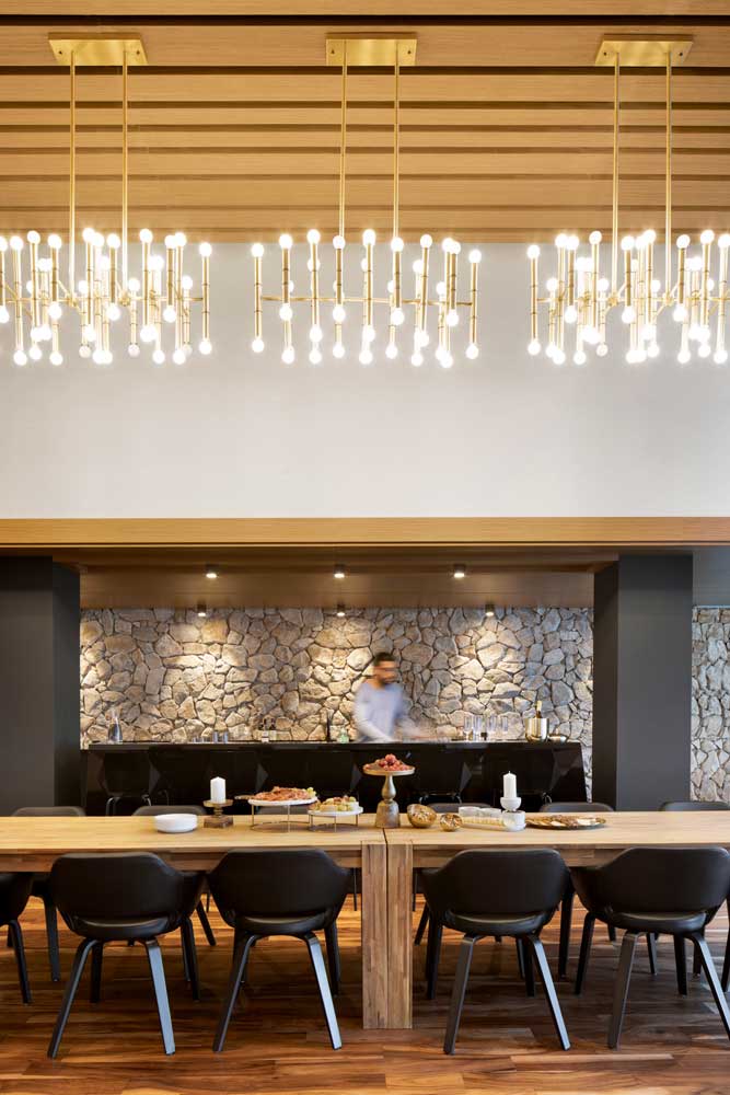 Party room dining table with 3 golden light fixtures above and the modern black bar behind. The backdrop behind the bar is a contrasting stone wall