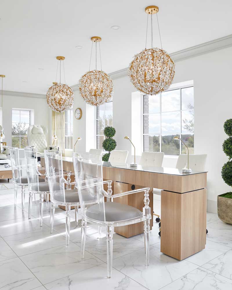 Long manicure desk with multiple seating spaces and three large globe-shaped light fixtures with nature inspired motifs facing beautiful outdoor views through the windows