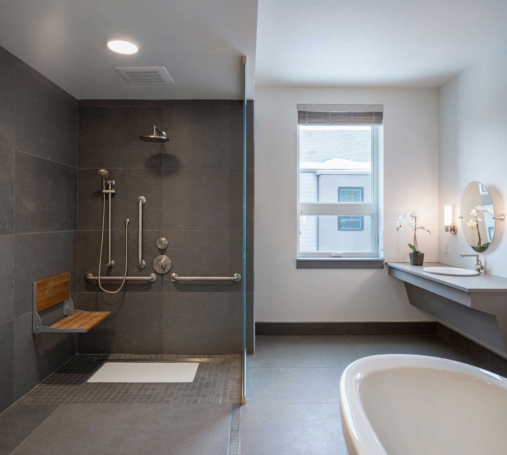 Accessible bathroom with a curbless shower equipped with a bench and grab bars, free standing tub and barrier free vanity. The colour scheme is predominantly grey with white walls around the vanity