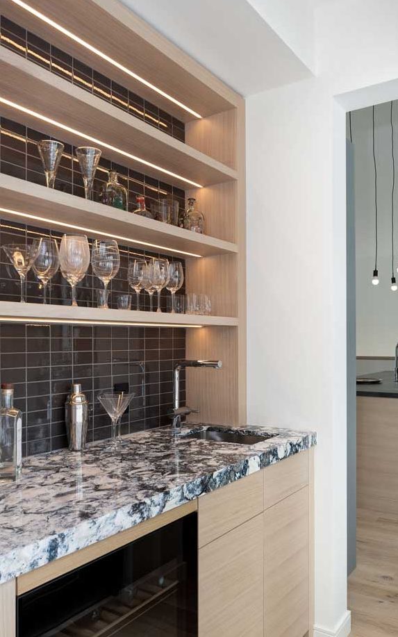 Wet bar just off the kitchen area done in light wood, dark wall tile behind the shelving and a bold-patterned countertop