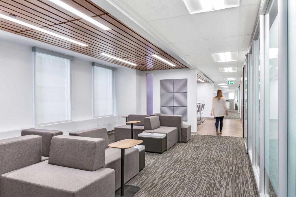 A woman walks through an interior hallway with taupe seating and repeated wood slats on the ceiling.