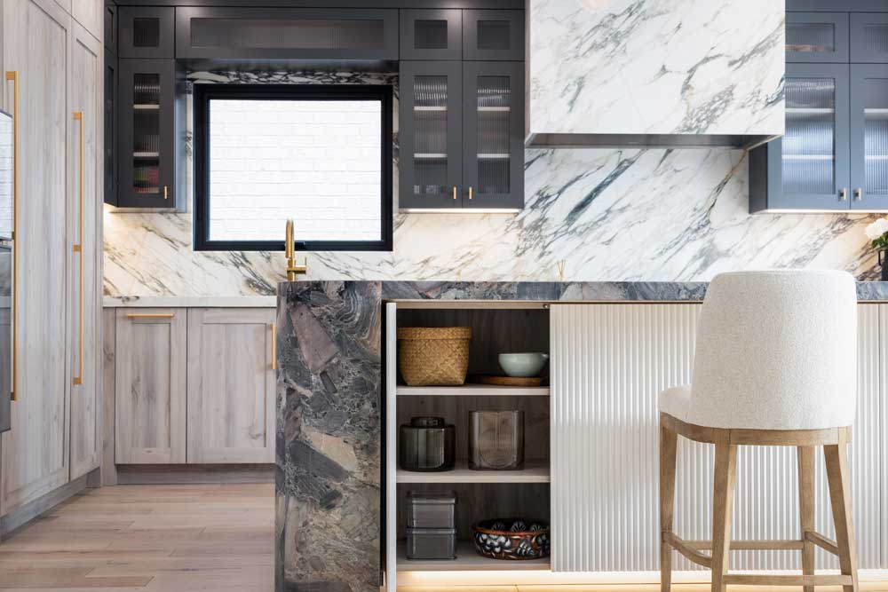 Kitchen countertops and backsplash are covered in luxurious marble with black upper cabinets and light wood finish on the bottom cabinetry. Island covered in a darker coloured marble and a storage cabinet underneath