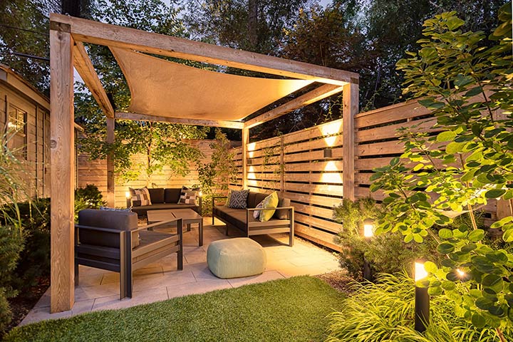 Inviting backyard with a wooden tall privacy fence all around, beautiful modern lighting, and modern black furniture under a wooden pergola.