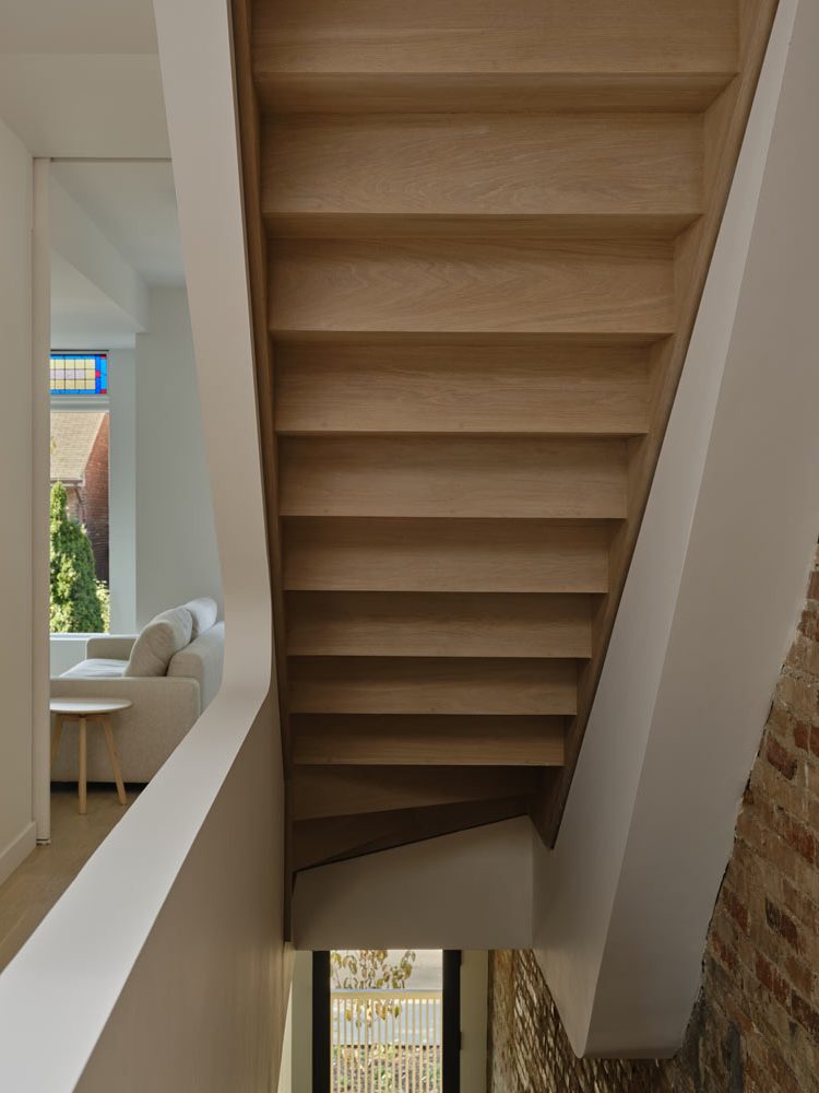 Underside of the wood finished staircase in contrast to the surrounding white walls and a brick wall visible on one side under the stairs