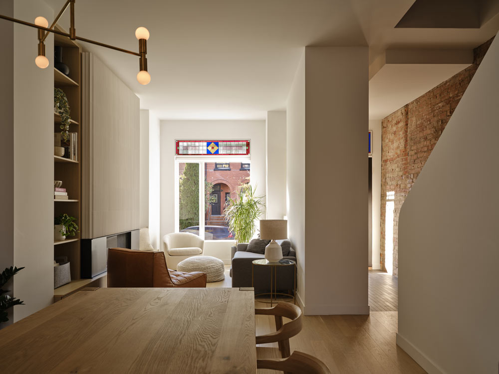 A view of the living room from the dining area, all is in predominantly white colours scheme with some warm wood details, brass light fixture and leather furniture pieces