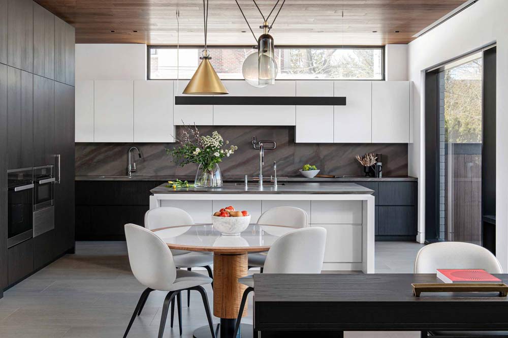 Modern duo tone kitchen with white upper cabinets and black on the bottom, with a drak marble patterned stone on the backsplash. There is a small round table in front of the small island