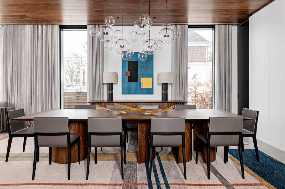 Dining room in mid century modern style. The focal point in the room is a blue modern art work on the wall between two windows. Light fixture made of multiple glass ball pendants hangs above a wooden dining table with upholstered chairs