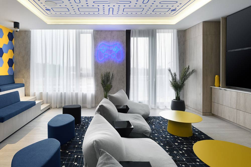 Gaming room in a monochromatic base with yellow and blue accents like the small coffee tables and seating against the wall. There are fun details like the blue LED neon gaming controller on the wall and a Pac Man like pattern on the ceiling.