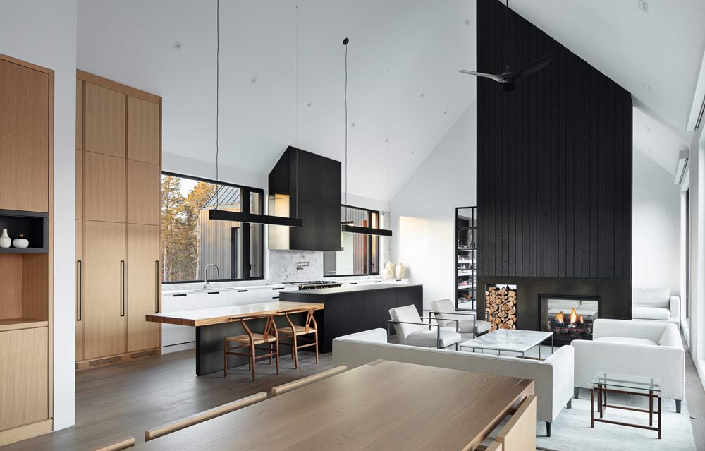 A view of the living area and kitchen in the background. Straight modern lines and wood and black finishes contrast the white slanted ceiling