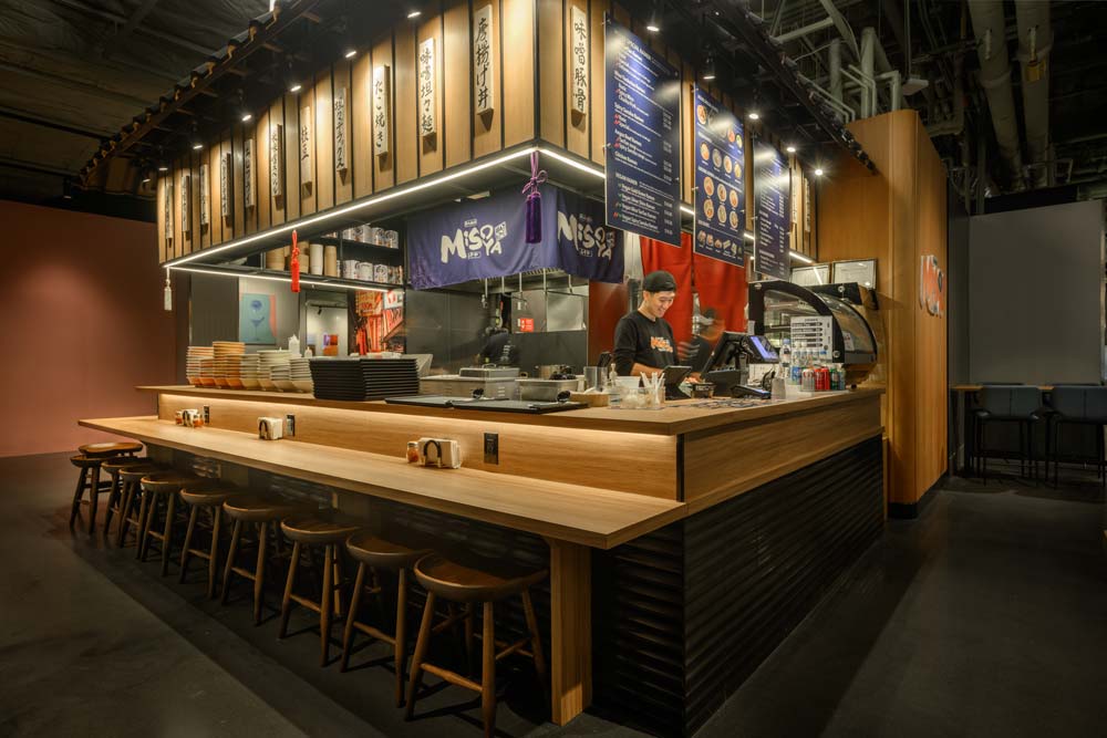 Environmental graphics are integrated in the wooden surround to detail the ramen shop experience, alongside the use of iconic Japanese elements such as the "Noren" fabric divider. Wooden countertop enhances the ambiance