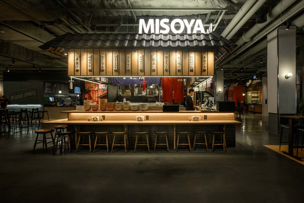 A view of the whole kiosk featuring wooden countertops all around the cooking area, and the black 'roof/ above displaying the name 'Misoya' in white lettering